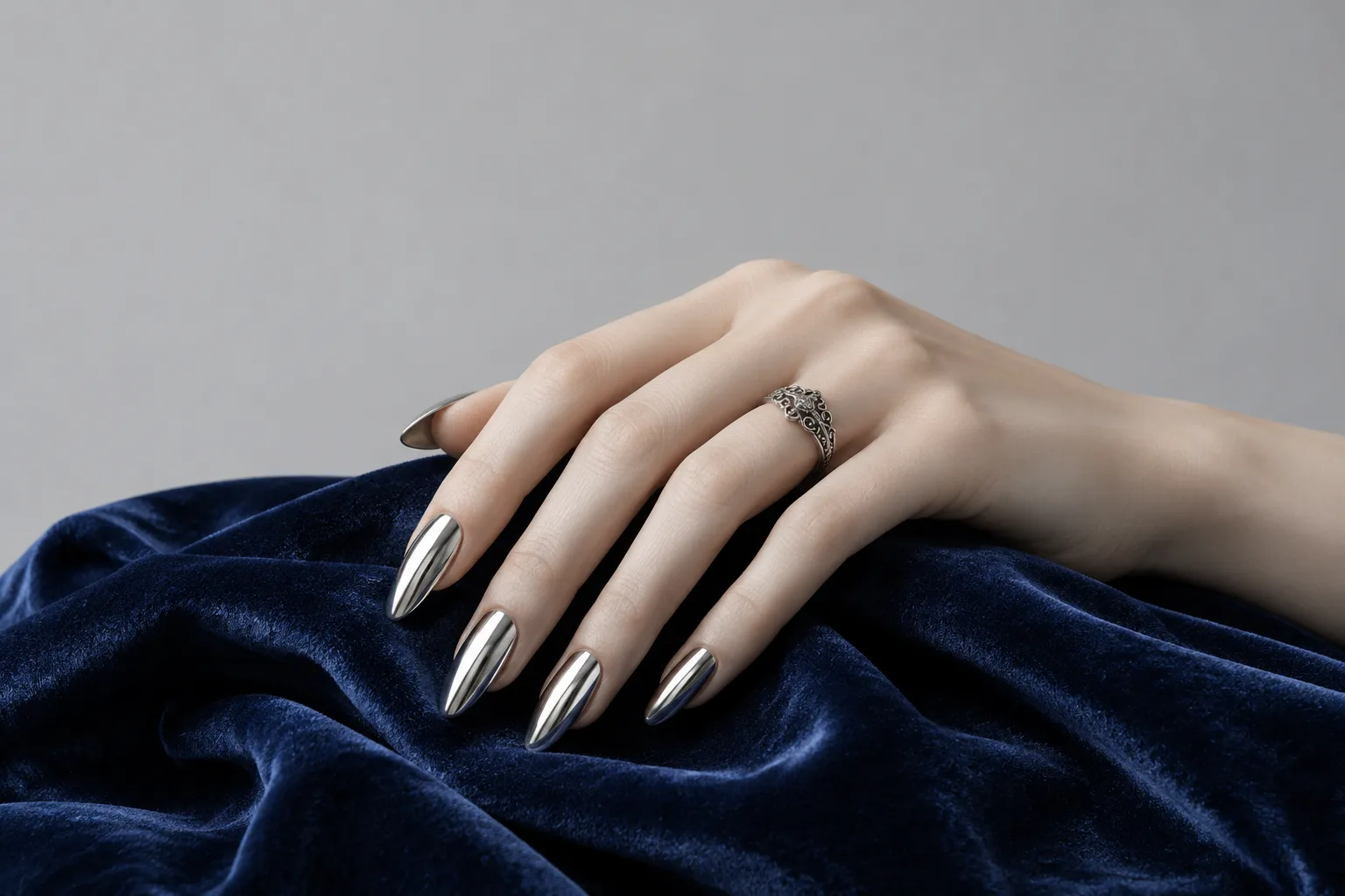 A pale hand with long chrome almond nails on navy velvet